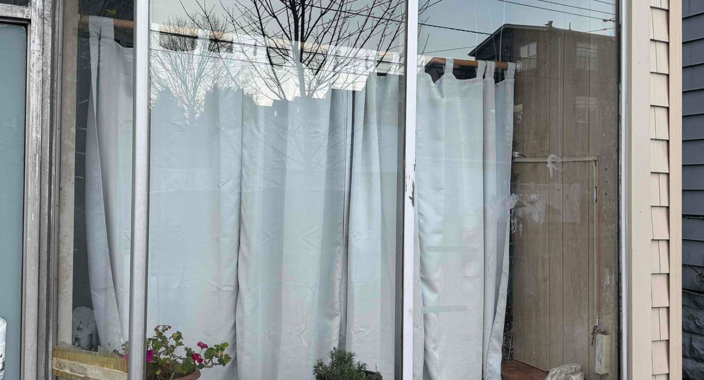 Exterior view of a large residential aluminum-framed window with white curtains visible inside, showing glass condition prior to repair service